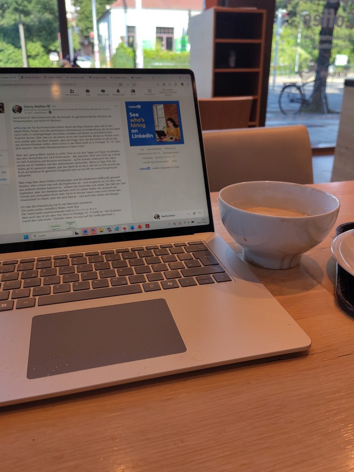 Foto: Laptop mit dem geöffneten LinkedIn-Beitrag zur Buchmarkt-Studie auf einem Holztisch in einer Bäckerei, daneben eine Schale Milchkaffee.