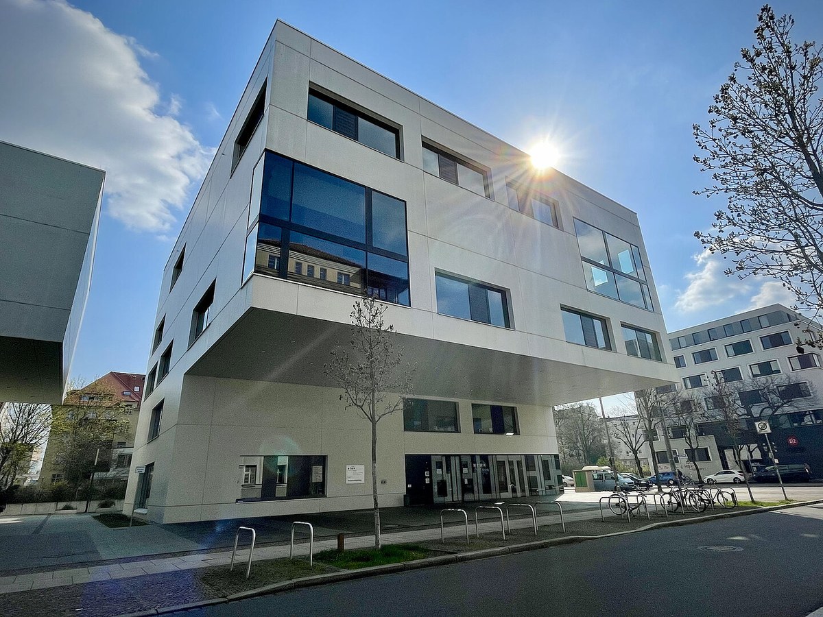 Gebäude der HTWK Leipzig: heller, kubischer Bau mit auskragendem Obergeschoss, dahinter blauer Himmel mit Sonne.