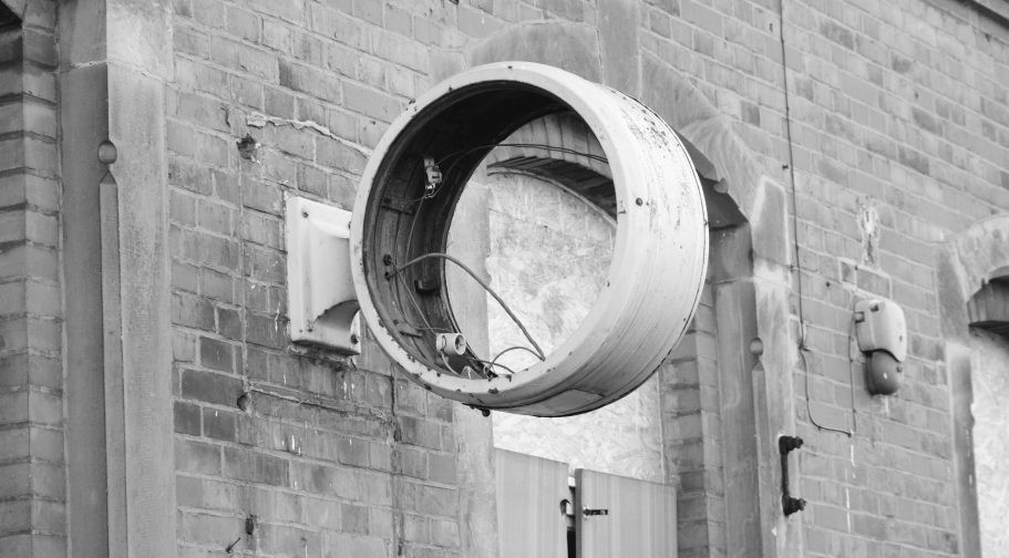 Schwarz-weißes Foto: ein runder, leerer Lampen- oder Leuchtengehäuse-Ring mit herausragenden Kabeln, der an einer Backsteinwand hängt; die Fassung ist leer, das Licht fehlt.