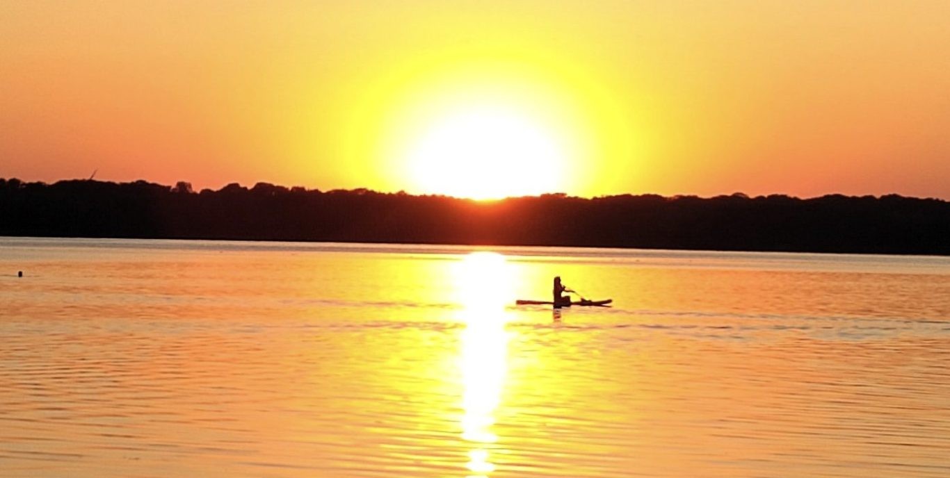 Sonnenuntergang über einem See: Die tiefstehende Sonne spiegelt sich in einer gelb-orangen Lichtbahn auf dem Wasser, davor die Silhouette einer Person auf einem Stand-up-Paddleboard, im Hintergrund ein dunkler Uferstreifen.