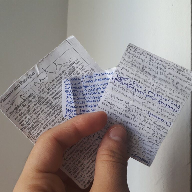 Foto: eine Hand hält mehrere kleine, eng beschriebene Zettel, darunter ein Zeitungsausschnitt „Vladimir and Estragon“ zu Samuel Becketts „Waiting for Godot“ und Zettel mit handschriftlichen Notizen zum Stück – klassische Spickzettel.